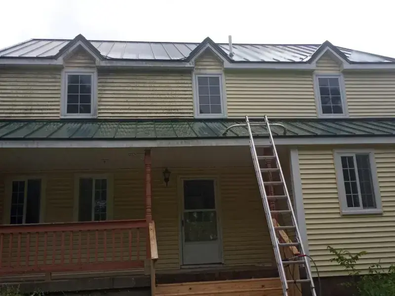 A large farmhouse with algae-streaked cream siding and a ladder standing against it, before cleaning